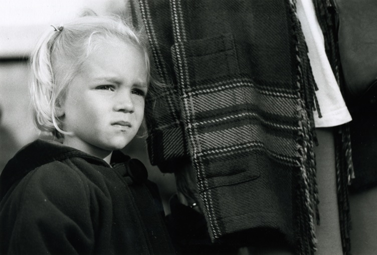A very blond little girl standing next to her mom. Mom is wearing a checked coat.