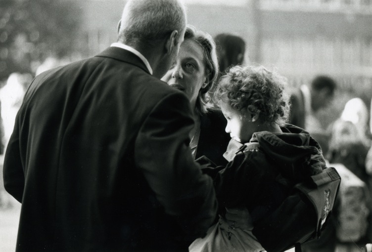 A curly haired boy - his face lit up by the sun - sitting on the arm of his mum. His mum is talking to a gentleman with his back to the camera.