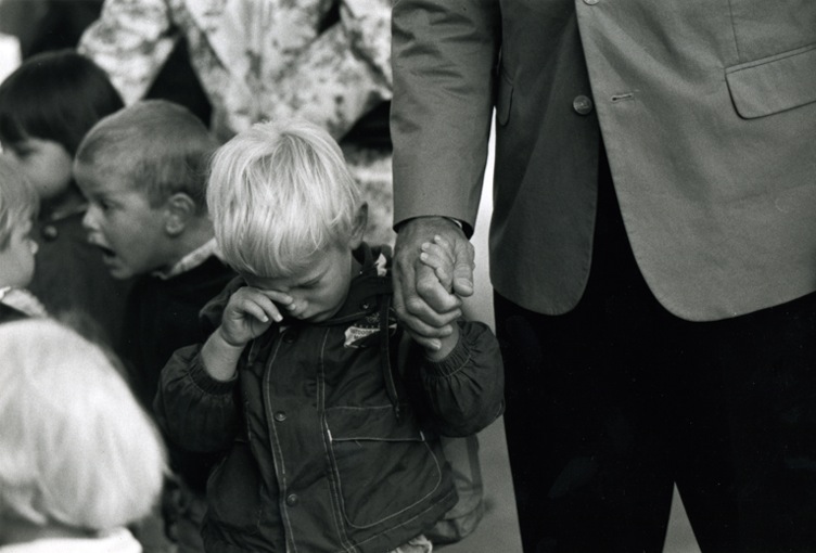 A little boy wiping a tear from his eye while holding the hand from his father. In the background another boy is shouting exictedly into the face of another child.